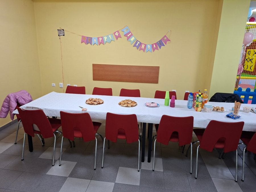 Маса за възрастни с червени столове, гирлянд Happy Birthday и детска площадка вдясно