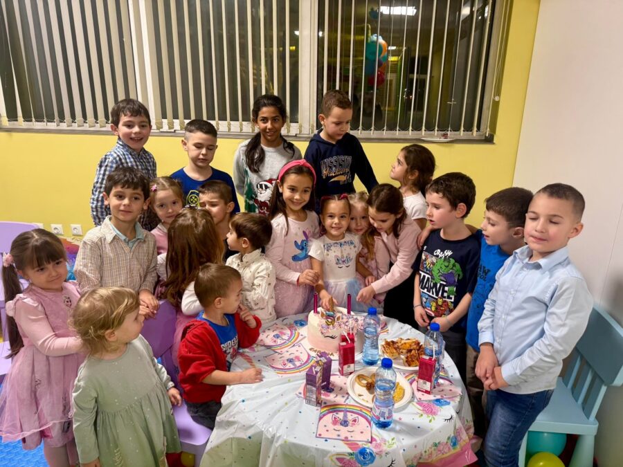 Много деца около маса с покривка на еднорози, бяла торта и бутилки с вода
