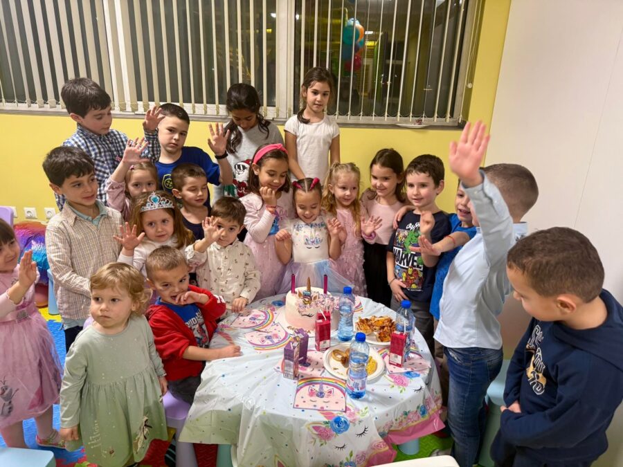 Торта с рог на еднорог и свещички, около нея деца вдигат ръце