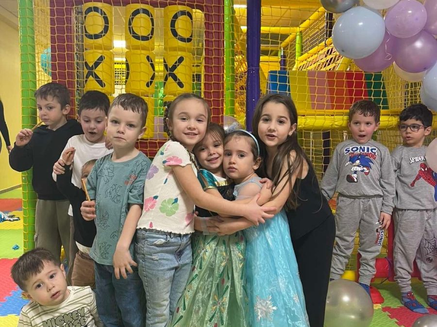 Група деца се прегръщат пред цветната игрална конструкция под лилаво-бял балонен свод