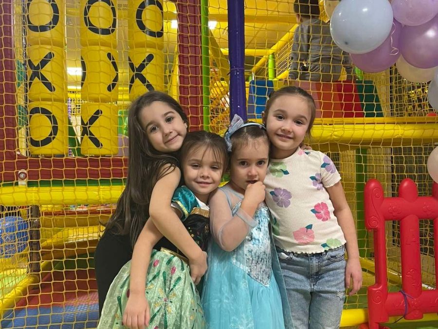 Четири момичета в рокли се прегръщат пред жълтата игрална конструкция и балони