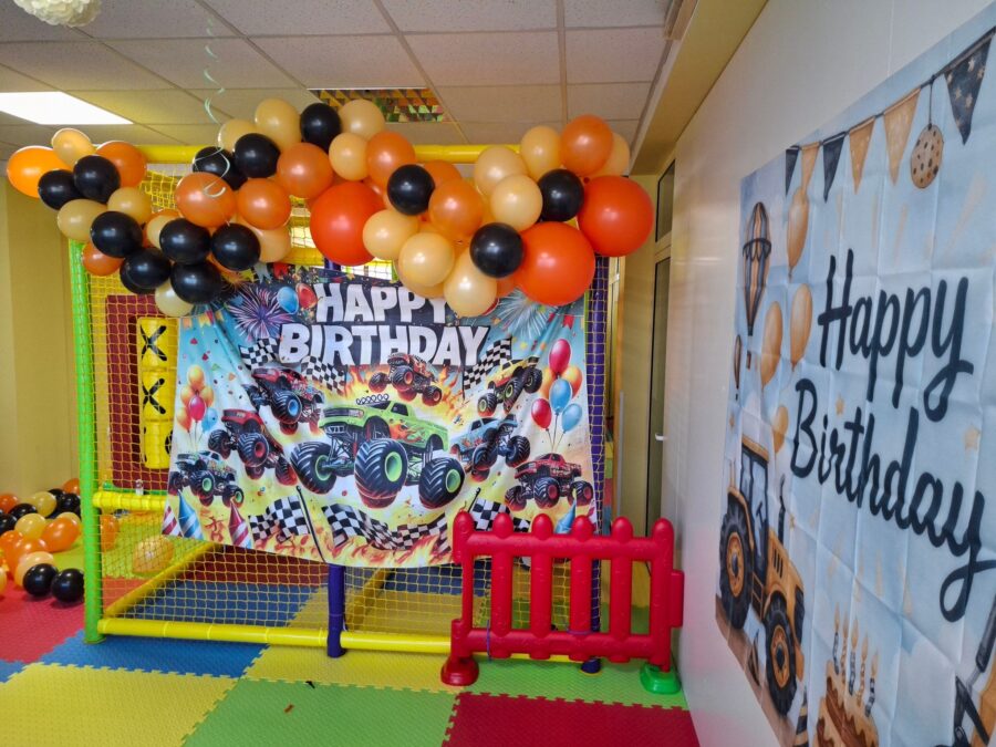 Фон с монстър трак камиони и надпис Happy Birthday, арка от оранжеви балони и червена оградка