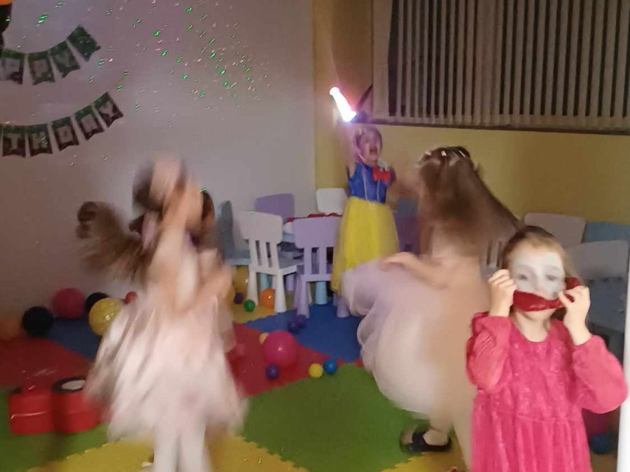 Деца танцуват под диско светлини в цветната зала, а момиченце в розова рокля се снима вдясно