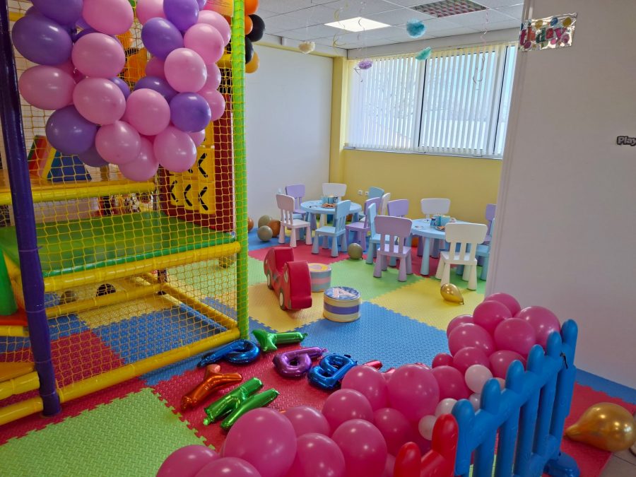 Двуетажна игрална конструкция с лилави и розови балони, синя ограда и детски столчета върху цветен под